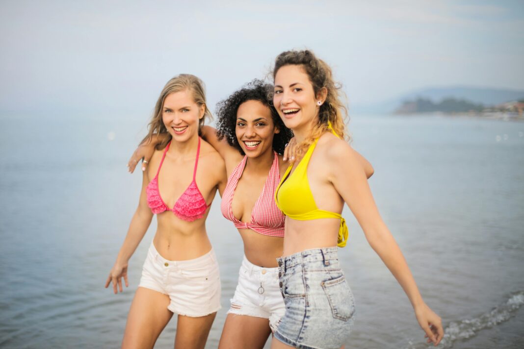 Drei junge Frauen in Shorts und Bikinioberteilen am Strand. Sie lachen