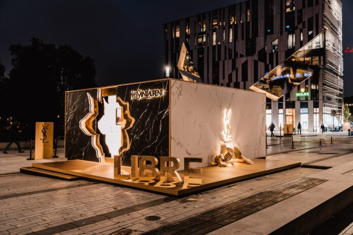 Aufnahme bei Nacht. In Braun- und Goldtönen gestalteter Container, abgestellt auf einem Platz mit einer HÄuserfassade im Hintergrund.